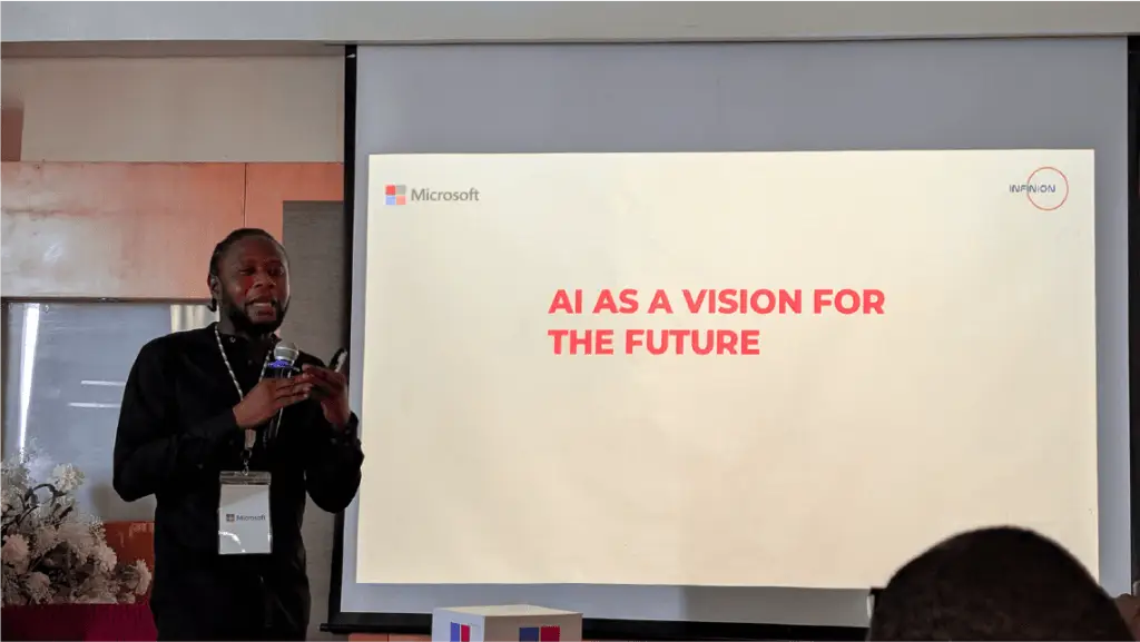 Speaker presenting on a stage beside a projector screen reading 'AI AS A VISION FOR THE FUTURE' with a Microsoft logo, microphone in hand and conference badge visible.