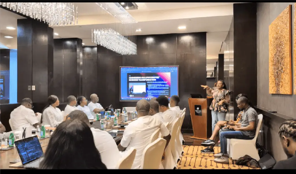 Presenter at a podium addresses a conference room filled with professionals around a long table; slides are on a large screen behind.