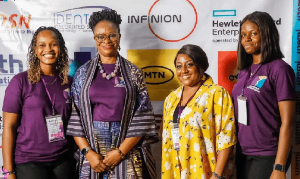 Four women stand together at a conference or expo, smiling at the camera; three wear purple shirts with name badges, one wears a yellow floral dress, banner with sponsor logos behind them.