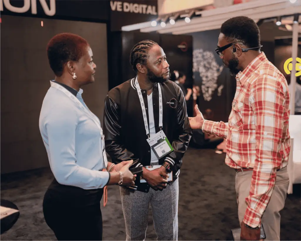 Three adults wearing conference badges converse in an exhibit hall at a busy event, center man with braided hair in a black jacket flanked by a woman and a man in a plaid shirt.