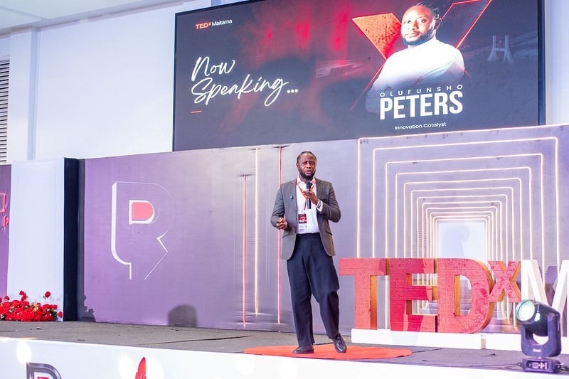 A man in a suit giving a TEDx talk on a stage with a large screen behind him that reads Now Speaking and features graphics and the TEDx logo on the right edge.